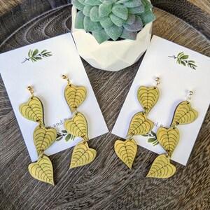 May include: Two pairs of gold-colored leaf-shaped earrings on white cards, displayed on a wooden tray. Each earring features multiple leaf pendants with detailed vein patterns. A small succulent plant in a white geometric pot is in the background.