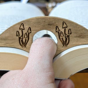 May include: A wooden bookmark shaped like a crescent moon with a design of mushrooms on each side. The bookmark is holding open a book.