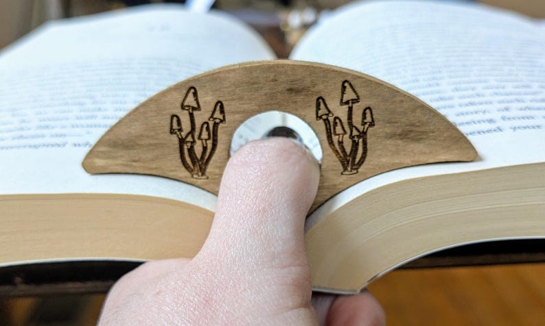 May include: A wooden bookmark shaped like a crescent moon with a design of mushrooms on each side. The bookmark is holding open a book.