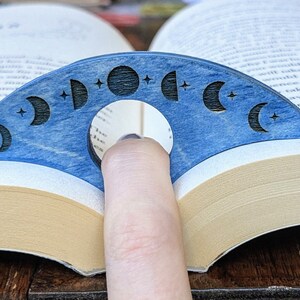 May include: A blue wooden bookmark shaped like a crescent moon with a cutout in the center for a finger. The bookmark is decorated with eight different phases of the moon and stars.