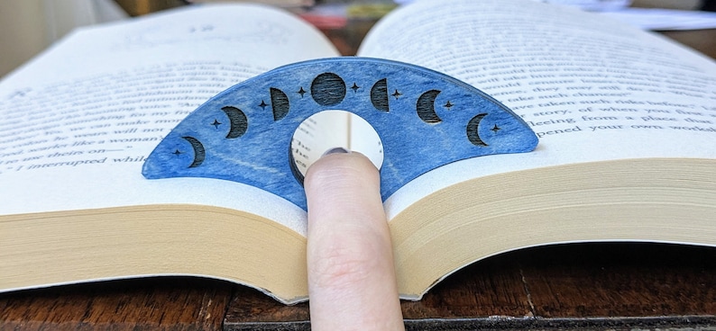 May include: A blue wooden bookmark shaped like a crescent moon with a cutout in the center for a finger. The bookmark is decorated with eight different phases of the moon and stars.