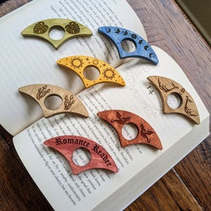 May include: Five different wooden bookmark designs with various engravings. The bookmarks are shaped like a crescent moon and feature designs such as a sun, a moon, a dragon, a mushroom, and a monstera leaf.