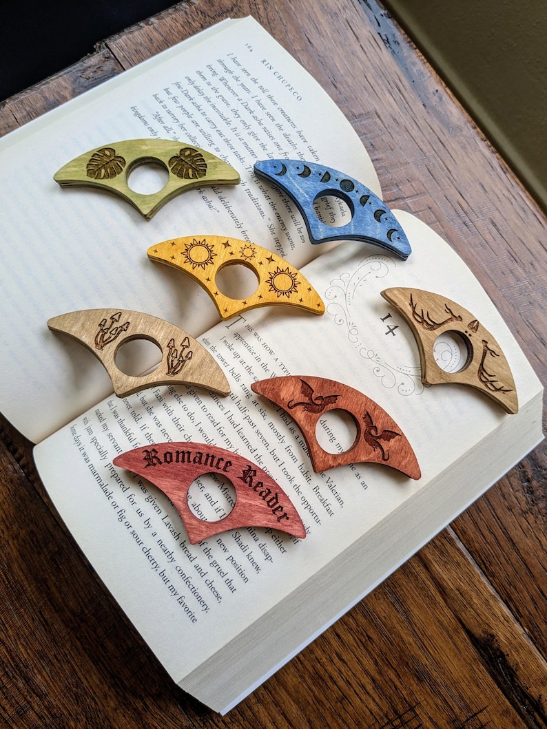 May include: Five different wooden bookmark designs with various engravings. The bookmarks are shaped like a crescent moon and feature designs such as a sun, a moon, a dragon, a mushroom, and a monstera leaf.