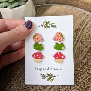 May include: A white card displays six wooden earrings with mushroom and frog designs. The earrings are painted in pink, red, green, and white. The card has the text "Stag and Branch" and is held by a person.