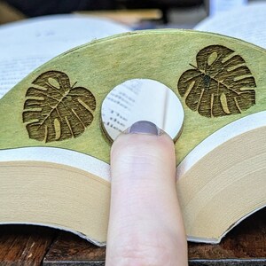 May include: A green wooden bookmark with two carved monstera leaves. The bookmark is shaped like a crescent moon and has a hole in the center for a finger.