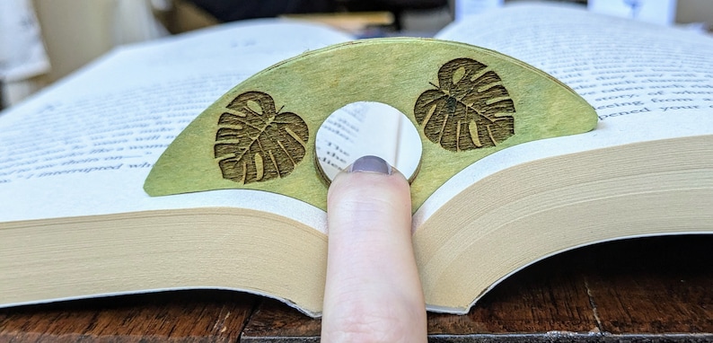May include: A green wooden bookmark with two carved monstera leaves. The bookmark is shaped like a crescent moon and has a hole in the center for a finger.