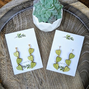 May include: Two pairs of wooden earrings with a cascading heart design. Each earring features four green wooden hearts connected by a silver or gold wire. The earrings are displayed on white cards with the text "Stag and Branch" printed on them.