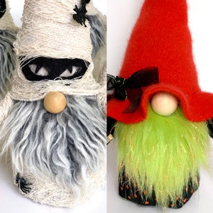 Op de afbeelding: Halloween kabouter decoraties. Eén kabouter is gewikkeld in crèmekleurige stof met een zwart gezicht en een houten kralen neus. De andere kabouter heeft een rode hoed, groene baard en een houten kralen neus. Beide kabouters hebben een zwarte strik.