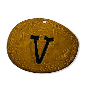Peut inclure: Une étiquette ovale en métal de couleur dorée avec une lettre V noire au centre. L'étiquette a une bordure décorative de petits triangles.