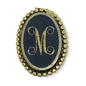 Może przedstawiać: Złota owalna broszka z czarnym emaliowanym tłem i złotą literą "M" w ozdobnym stylu.