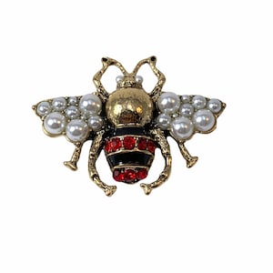 Puede incluir: Broche de abeja dorada con alas de perlas y cuerpo rojo y negro. El broche presenta una cabeza y patas doradas, con las alas compuestas por numerosas perlas pequeñas y redondas. El cuerpo tiene una banda negra con gemas rojas.