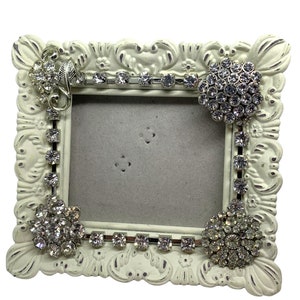 May include: A white ornate picture frame with rhinestones and a gray background. The frame has a decorative design and is embellished with sparkling rhinestones.