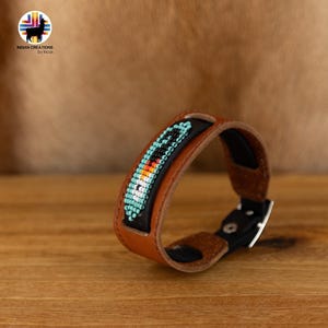 Può includere: Un braccialetto in pelle marrone con un design di perline nere che include perline turchesi, bianche, arancioni e nere. Il braccialetto ha una chiusura a fibbia nera.