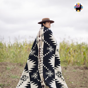 May include: A person wearing a black and white patterned blanket with a brown hat. The blanket has a geometric design with black and white triangles and squares. The person is standing in a field with tall grass.