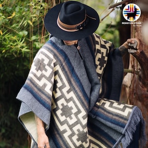 May include: A gray and white patterned poncho with fringe, worn by a person wearing a black fedora and blue jeans. The poncho has a geometric design with a brown and blue border.