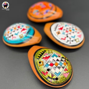 May include: Four hand-painted, oval-shaped ocarinas in shades of orange and brown. Each ocarina features a unique geometric design in various colors, including blue, red, yellow, and white. The ocarinas are displayed on a dark gray surface.