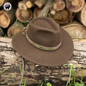 Może przedstawiać: Brązowy kapelusz typu fedora z ozdobną opaską z kolorowym geometrycznym wzorem. Kapelusz ma szerokie rondo i klasyczną koronę. Kapelusz spoczywa na drewnianej powierzchni z trawą.