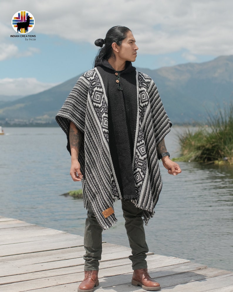 Pode incluir: Um poncho listrado preto e branco com padr&otilde;es geom&eacute;tricos. O poncho est&aacute; sendo usado por uma pessoa em p&eacute; em um cais de madeira. A pessoa est&aacute; vestindo uma camisa preta com capuz e cal&ccedil;as verde-escuras.