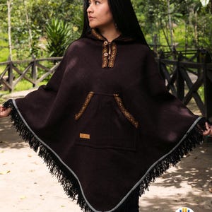 May include: A dark brown hooded poncho with a pocket and decorative trim. The poncho has a fringe along the bottom edge and is made of a thick, woven material. The trim is a lighter brown with a geometric pattern. The poncho is worn outdoors.