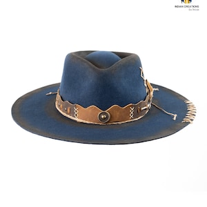 Op de afbeelding: Een blauwe wollen fedora hoed met een bruine leren band en decoratieve stiksels. De hoed heeft een brede rand en een verweerde look.