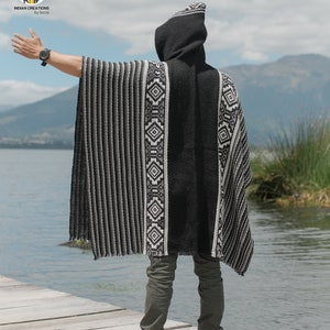 Pode incluir: Um poncho preto com capuz, listras brancas e cinzas e padr&otilde;es geom&eacute;tricos. O poncho est&aacute; sendo usado por uma pessoa em p&eacute; em um p&iacute;er de madeira com vista para um lago.