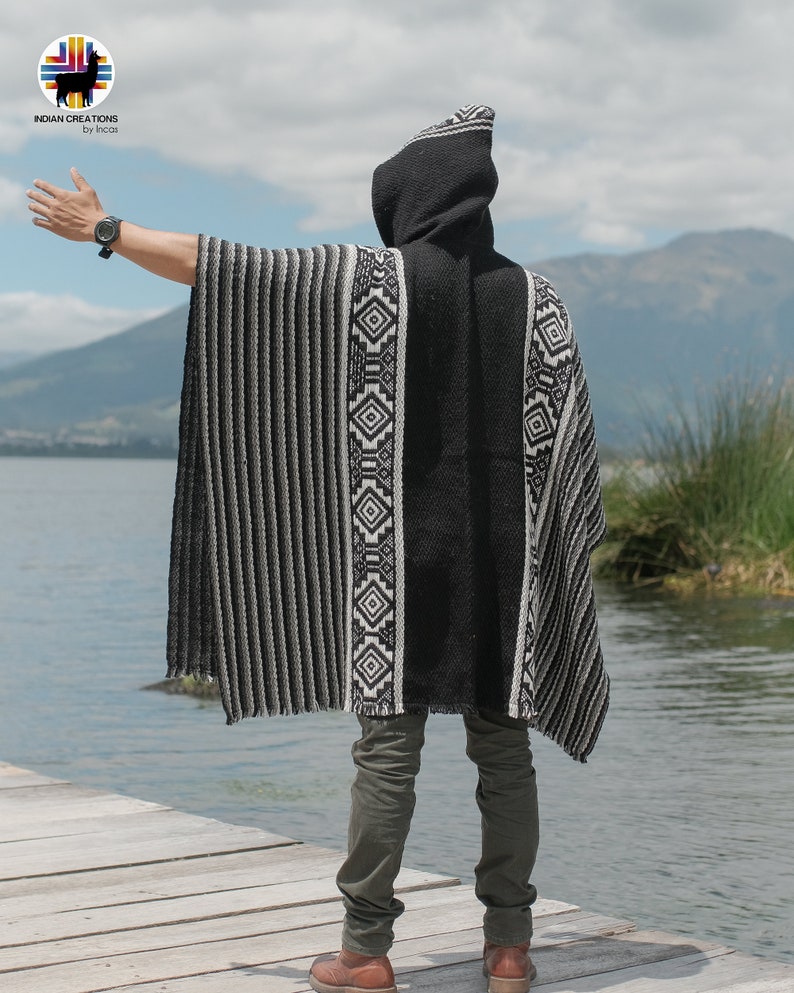 Pode incluir: Um poncho preto com capuz, listras brancas e cinzas e padr&otilde;es geom&eacute;tricos. O poncho est&aacute; sendo usado por uma pessoa em p&eacute; em um p&iacute;er de madeira com vista para um lago.