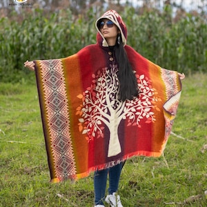 May include: A woman wearing a red, orange, and white hooded poncho with a tree design. The poncho has a fringe border and a button closure. The woman is standing in a field of green grass.