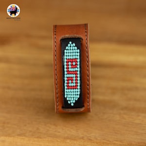 Può includere: Un braccialetto in pelle marrone con un design di perline turchesi e rosse. Il braccialetto è cucito con filo nero e ha un retro in pelle nera.