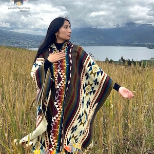 Puede incluir: Una mujer con un poncho con capucha de colores con patrones geométricos, de pie en un campo de hierba alta con un lago y montañas en el fondo.