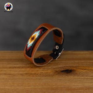 Può includere: Un braccialetto in pelle marrone con una fibbia nera e un design di perline colorate. Le perline sono disposte in un motivo geometrico con colori rosso, giallo, blu e bianco.