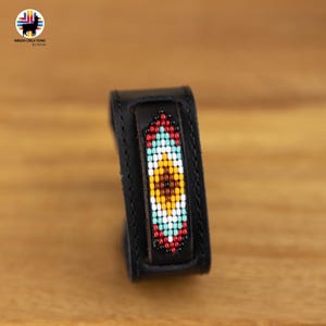 Può includere: Un braccialetto in pelle nera con un design di perline colorate. Le perline sono disposte in un motivo geometrico con colori rosso, giallo, blu e bianco.