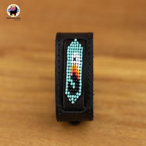 Può includere: Un braccialetto in pelle nera con un design di perline turchesi, bianche, arancioni e nere. Il design è una piuma stilizzata.