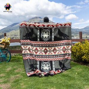 May include: A gray blanket with a wolf design. The blanket features a central wolf face and howling wolf silhouettes, with geometric patterns in red, white, and black. The blanket is displayed outdoors on a grassy area.