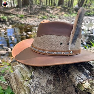 May include: A brown leather cowboy hat with a tan burlap band and a brown feather accent. The hat is sitting on a piece of weathered wood.
