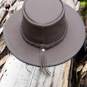 May include: A brown leather hat with a wide brim and a braided band. The hat features a decorative leather strap with a silver stud and fringe. The hat is a neutral brown color and appears to be made of high-quality leather.
