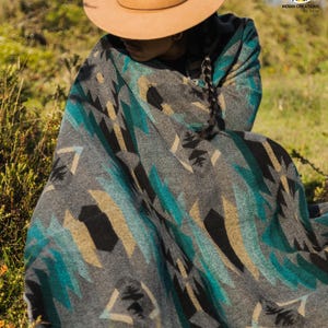 Könnte beinhalten: Eine graue Decke mit einem geometrischen Muster in Türkis, Schwarz und Beige. Die Decke ist über einer Person drapiert, die einen beigen Hut trägt. Das Design der Decke weist eckige Formen und stilisierte Motive auf, wodurch ein strukturiertes Aussehen entsteht. Die Decke ist für den Außenbereich geeignet.