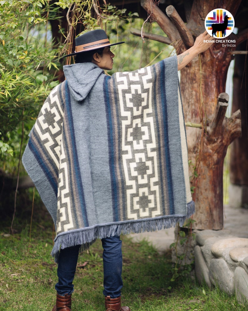 May include: A gray hooded poncho with a geometric pattern in white, brown, and blue. The poncho has a fringe border and is worn by a person wearing a brown hat and blue jeans.