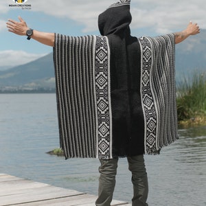 Pode incluir: Uma pessoa vestindo um poncho com capuz listrado preto e branco com padr&otilde;es geom&eacute;tricos. O poncho &eacute; feito de um material macio e quente e tem um caimento relaxado. A pessoa est&aacute; em p&eacute; em um p&iacute;er de madeira com vista para um corpo de &aacute;gua.