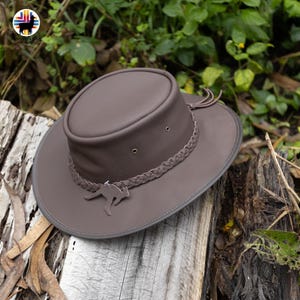 May include: A brown leather hat with a wide brim and a braided band. The hat features a kangaroo emblem and a decorative tassel. The hat is sitting on a wooden surface, with green foliage in the background.