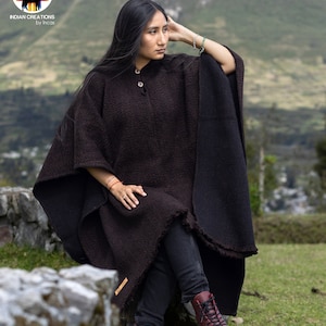 May include: A woman wearing a brown alpaca wool poncho with a black trim. The poncho has a button closure and is made in Peru. The woman is sitting on a rock with a mountain in the background.