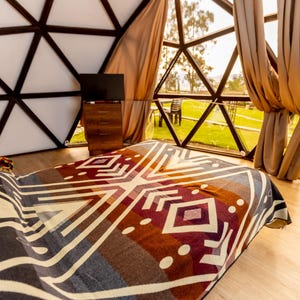 Op de afbeelding: Een bed met een patroon deken in een geodetische koepel. De deken heeft geometrische ontwerpen in grijstinten, bruin en bordeauxrood. Een houten dressoir en een tv zijn zichtbaar op de achtergrond.