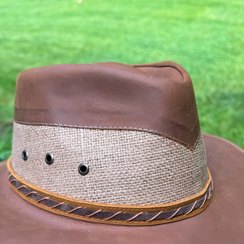 May include: A brown leather and woven straw hat with a wide brim and a leather band around the crown. The hat has three eyelets on the crown.