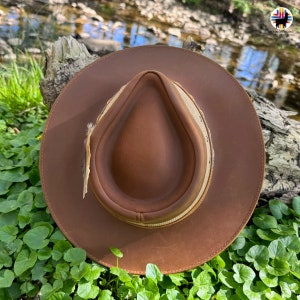 May include: A brown leather hat with a wide brim and a tan leather band. The hat is sitting on a bed of green leaves.