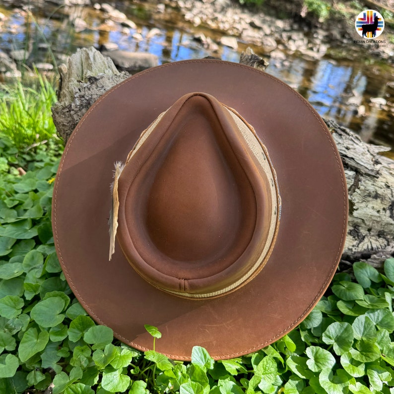 May include: A brown leather hat with a wide brim and a tan leather band. The hat is sitting on a bed of green leaves.