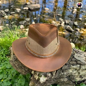 May include: A brown leather cowboy hat with a brown and tan woven band and a feather accent. The hat is sitting on a tree branch with green foliage in the background.