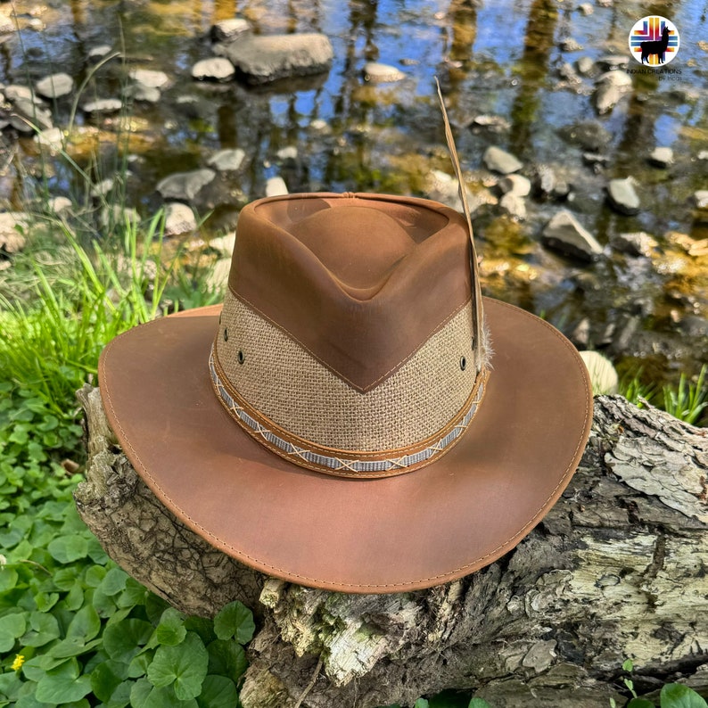 May include: A brown leather cowboy hat with a brown and tan woven band and a feather accent. The hat is sitting on a tree branch with green foliage in the background.