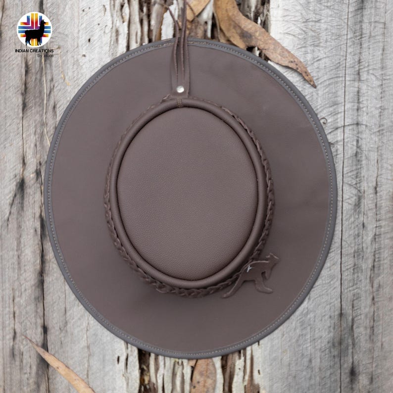 May include: A brown leather hat with a wide brim and a braided band around the crown. The hat features a kangaroo emblem and a leather strap. The hat is displayed against a wooden background.
