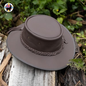 May include: A brown leather hat with a wide brim and a braided band around the crown. The hat has a round top and is decorated with small metal eyelets. The hat is sitting on a wooden surface.