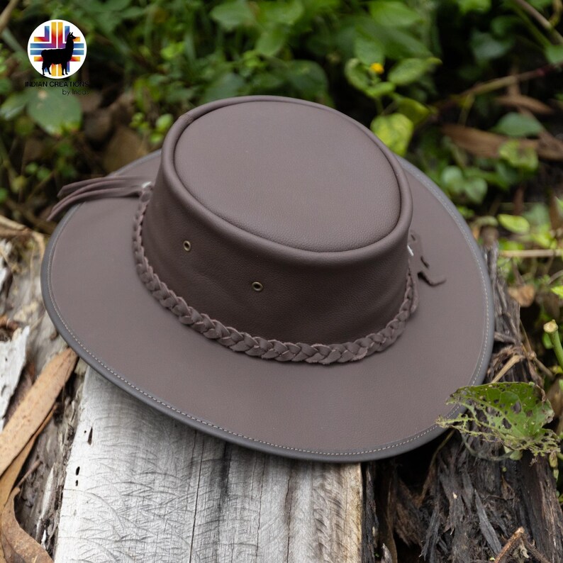 May include: A brown leather hat with a wide brim and a braided band around the crown. The hat has a round top and is decorated with small metal eyelets. The hat is sitting on a wooden surface.