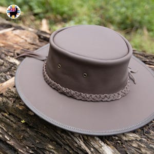 May include: A taupe-colored leather hat with a wide brim and a braided band. The hat features a rounded crown and decorative fringe. The brim has a gray trim. The hat is resting on a weathered log.